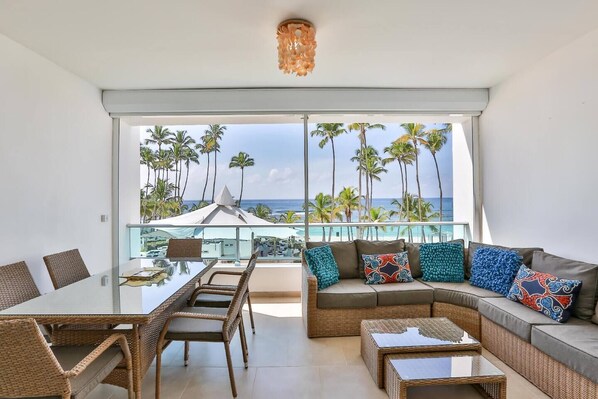 Ein geräumiger Balkon mit einem Glastisch und Korbmöbeln, mit Blick auf einen Strand mit Palmen und dem Meer.