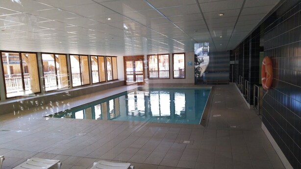Une piscine intérieure avec des parois en verre transparent, un sol carrelé et des chaises longues blanches.