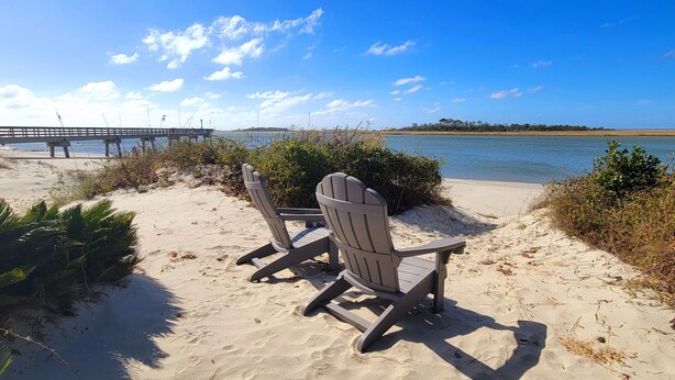 A beach with two Adirondack chairs facing the water, a wooden pier in the background, and a clear sky.