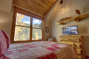 Un dormitorio con un ventanal grande, techo de madera, televisión y cama con colcha.