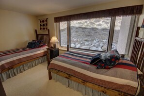 A bedroom with two beds, a nightstand with a lamp, and a large window offering a view of a rocky landscape. - Saints John 2526 (Keystone)