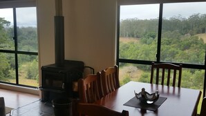 A dining area with a table and chairs, a wood-burning stove, and large windows offering a view of a forested landscape.