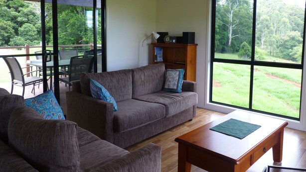 A living room with a sofa, a coffee table, and a dining area with chairs and a table. There is a large window providing a view of the outdoors.