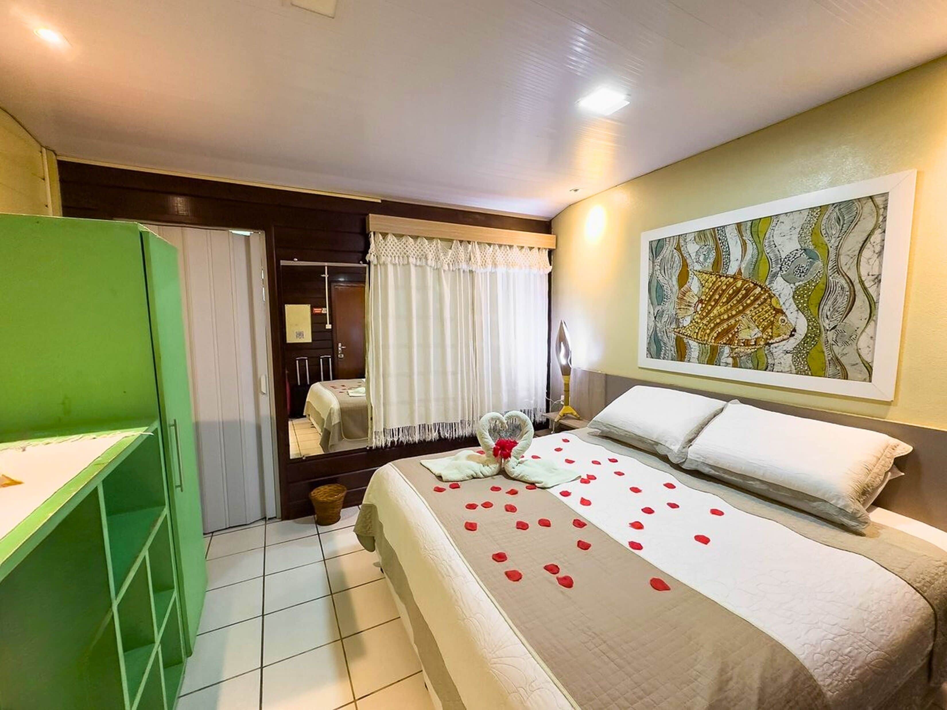 A hotel room with a bed, a green cabinet, a mirror, a painting, and a door leading to a bathroom.