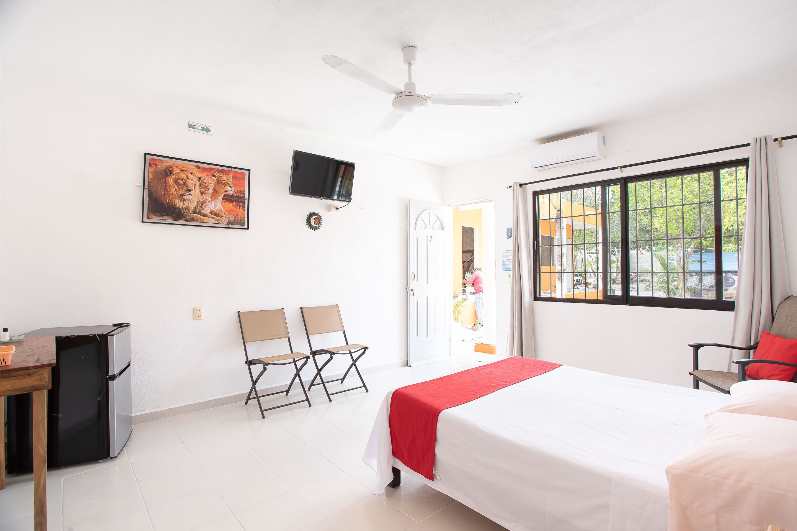 A hotel room with a bed, a desk, a refrigerator, two chairs, a television, and a window with a view of the outside.