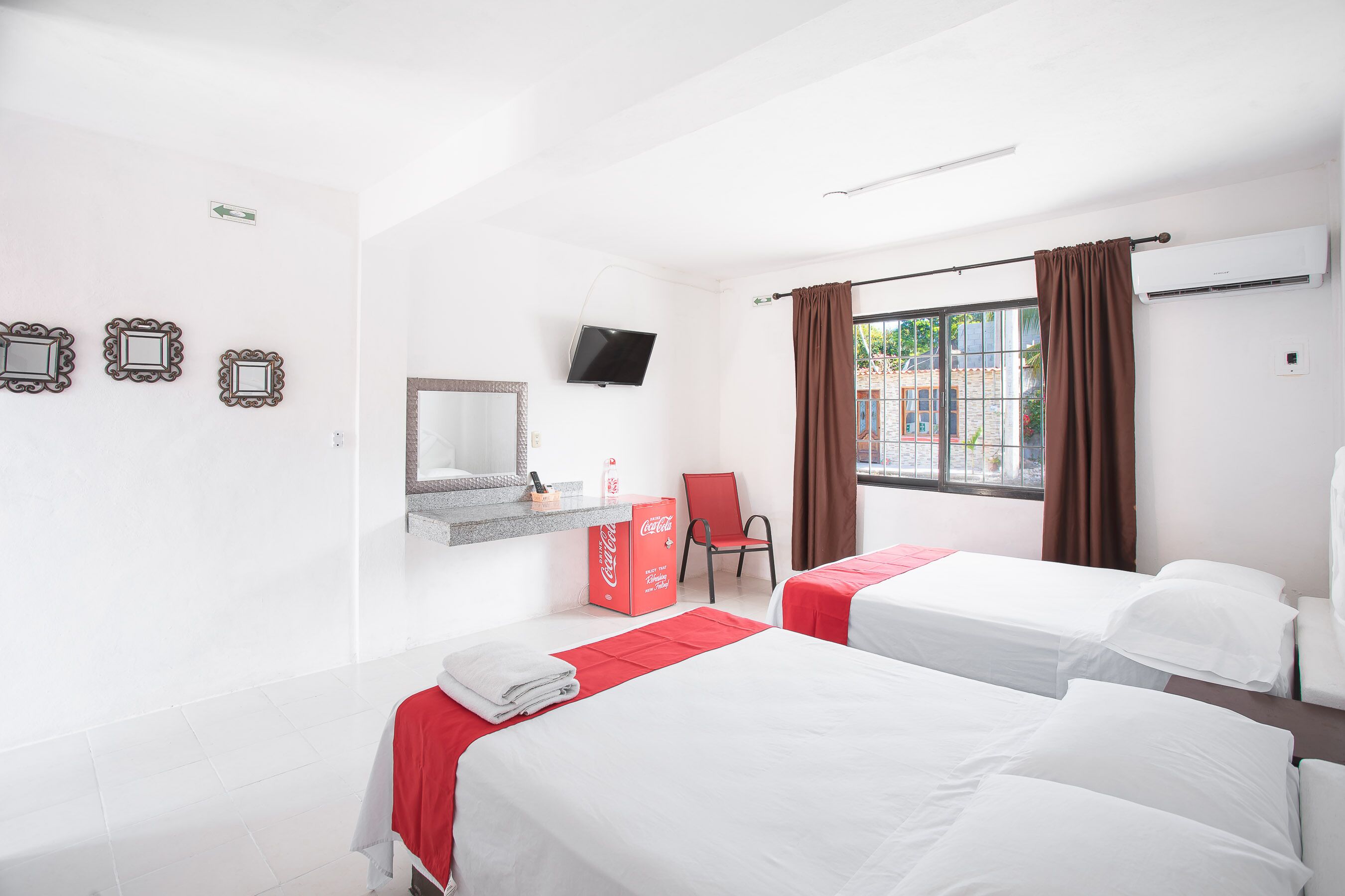A hotel room with two beds, a television, a red Coca-Cola cooler, a chair, and a window with curtains.