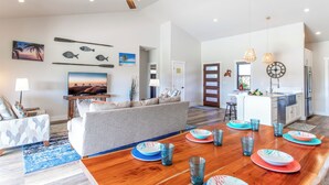 Amplia sala de estar con sofá, mesa de centro y televisor. Hay un comedor con mesa de madera y platos azules y anaranjados. La habitación tiene ventilador de techo y decoración mural.