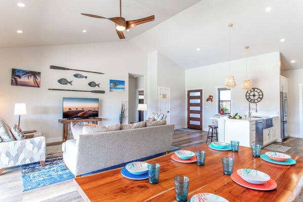 A spacious living room with a sofa, a coffee table, and a TV. There is a dining area with a wooden table and blue and orange plates. The room has a ceiling fan and wall art. - Ainamalu 3 Bdrm Home with Pool in Waikoloa Resort! (Waikoloa)