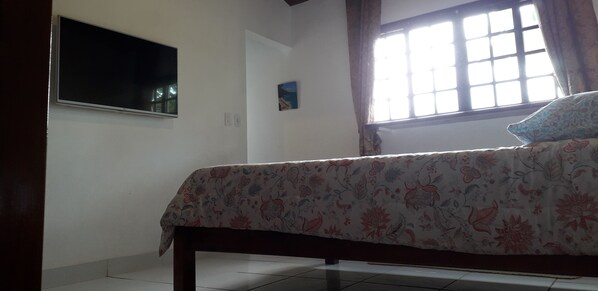 A bedroom with a bed, a TV mounted on the wall, a window with curtains, and a small painting on the wall.