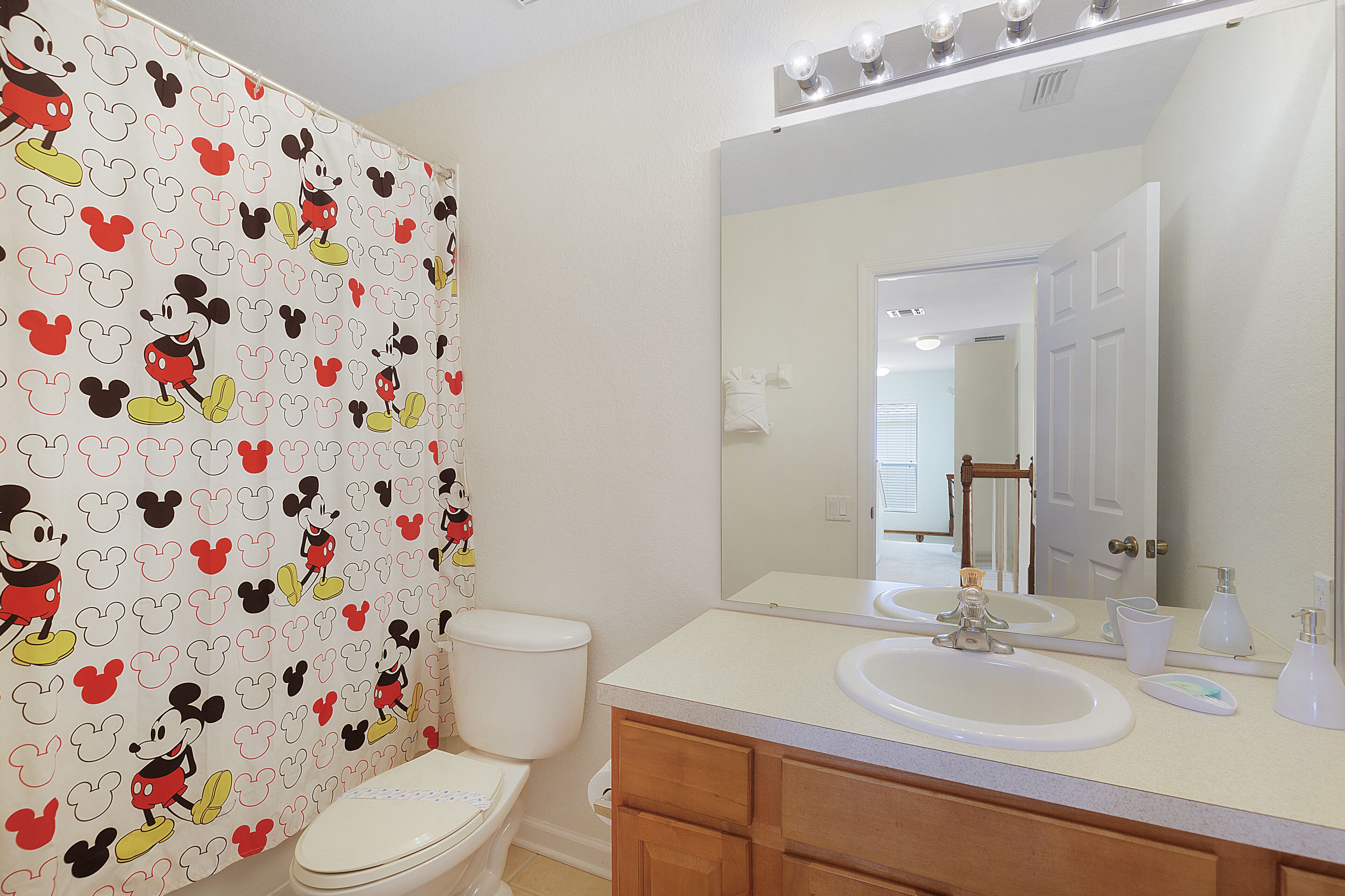Une salle de bain avec un rideau de douche à l’effigie de Mickey Mouse, des toilettes blanches, un lavabo avec un miroir au-dessus et des meubles en bois.