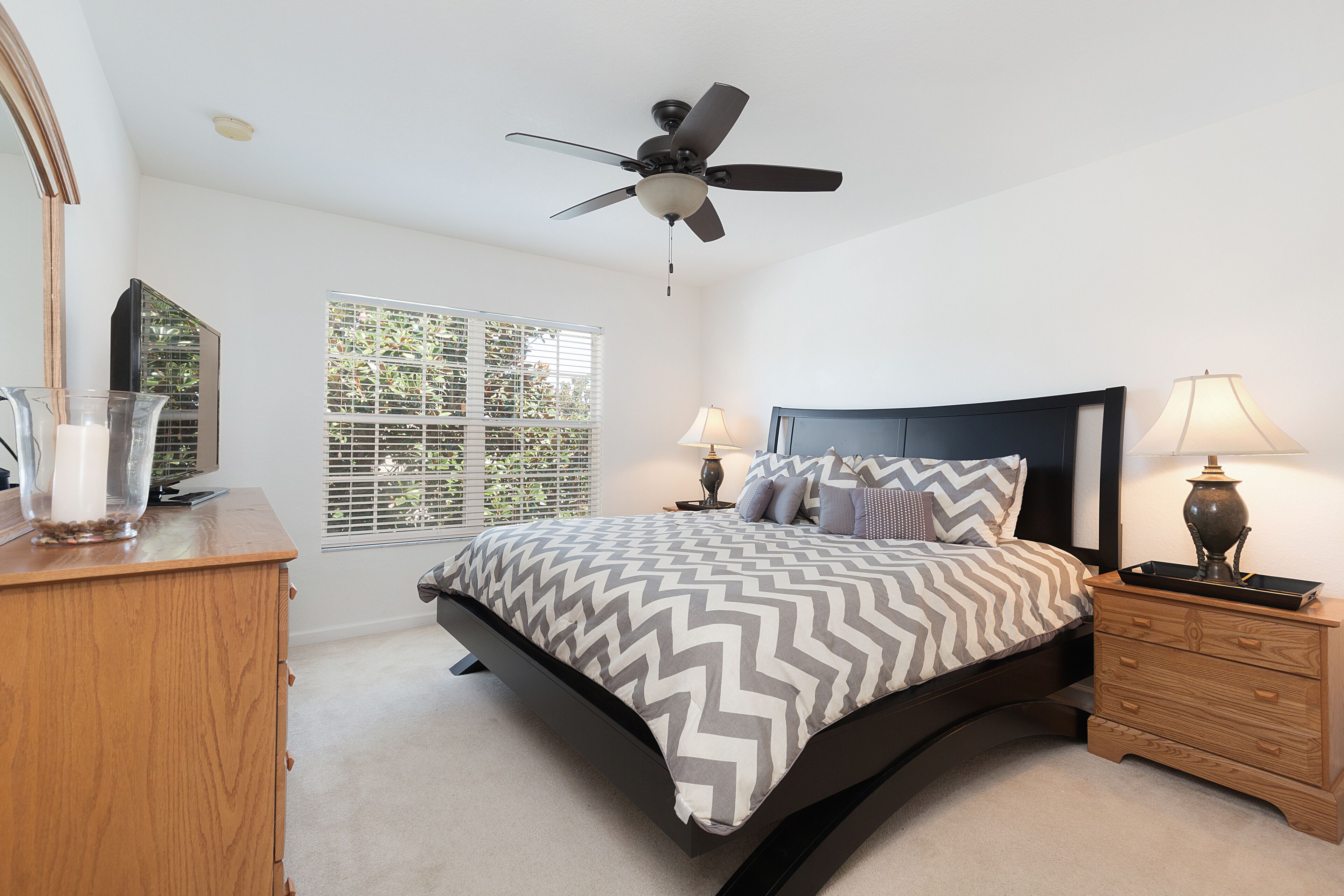 Une chambre à coucher comprenant un lit, des tables de chevet, une commode, un ventilateur de plafond et une fenêtre avec des stores.
