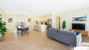 A modern living room with a grey sofa, wooden flooring, a kitchen area with a dining table, and a TV on a wooden stand. - Aldinga Beach Experience (Aldinga Beach)