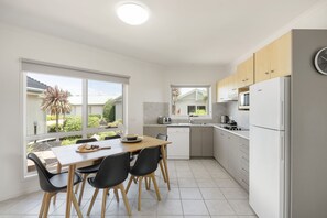 A modern kitchen with a dining area, white appliances, and large windows offering a view of the outdoors.