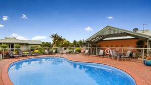 A swimming pool with lounge chairs and umbrellas, surrounded by a brick patio and palm trees.