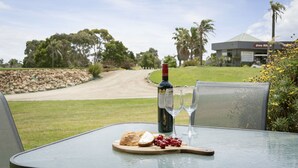 A table with a bottle of wine, two glasses, a plate with grapes and bread, and a view of a golf course and a building in the background.