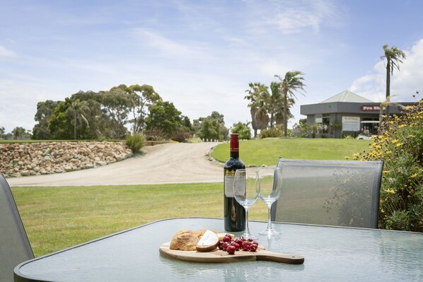 A table with a bottle of wine, two glasses, a plate with grapes and bread, and a view of a golf course and a building in the background.
