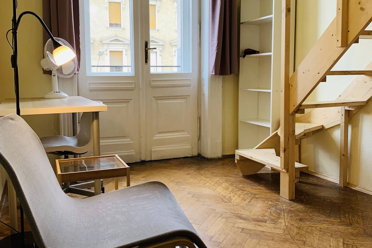 Una habitación con una silla de madera, una mesa, una lámpara de pie, un ventanal con cortinas y una escalera de madera.