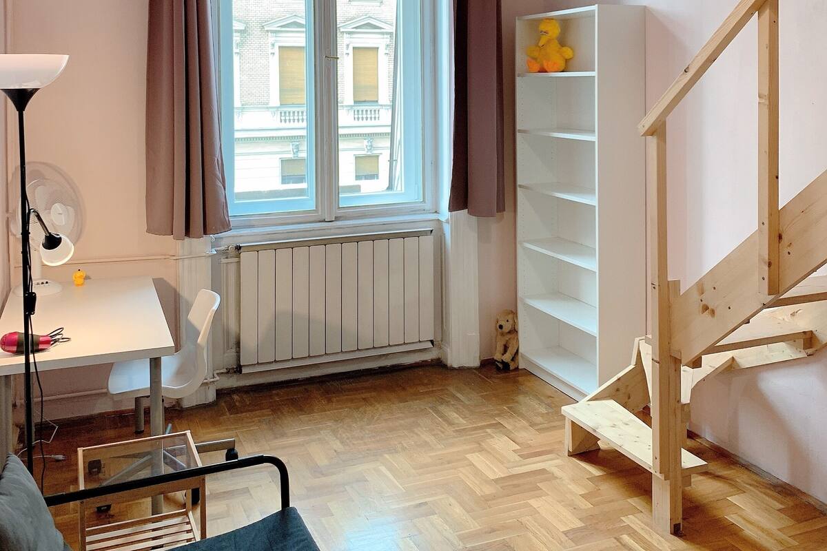 Una habitación con piso de madera, un escritorio con lámpara, una silla, una cama, una ventana con cortinas y una escalera de madera.