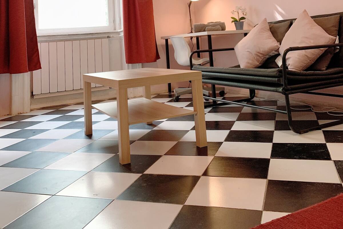 Una habitación con piso a cuadros, una cama con almohadas, una mesa, un ventanal con cortinas y una lámpara.