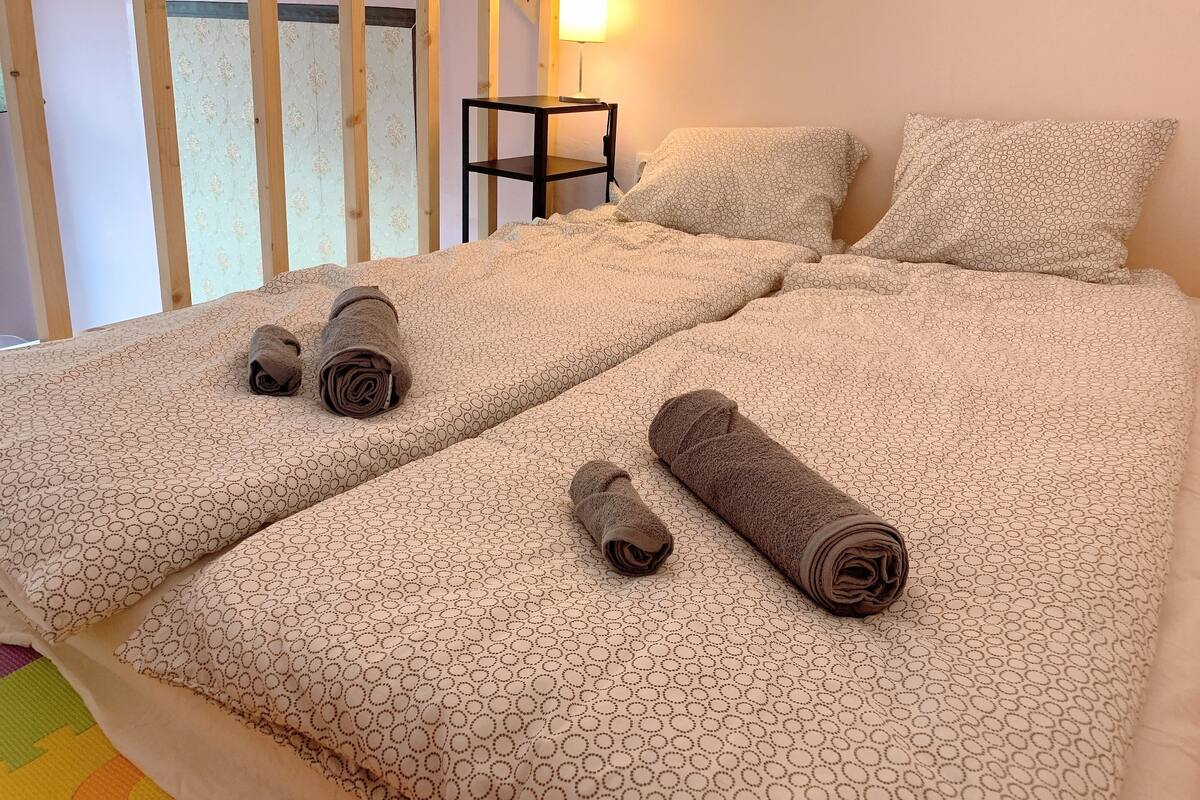 Una cama doble con una colcha de estampado geométrico, dos toallas enrolladas, una mesita de noche con una lámpara y almohadas estampadas.