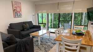 Una sala de estar moderna con un sofá gris, una mesa de comedor de madera con sillas blancas y un ventanal con vistas a zonas verdes.