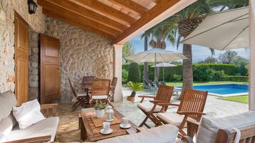 Un área de estar al aire libre techada, con paredes de piedra, muebles de madera y vistas a una piscina.