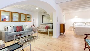 A spacious living room with a grey sofa, a small round table, and a wooden coffee table. There is a white bed with a wooden headboard in the background. - Genteel Home Catedral Sevilla (Sevilla)