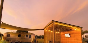 En træhytte med et fjernsyn, et overdækket udeområde med lyskæder, og en campingvogn på en græsplæne.