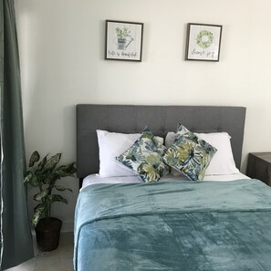 A bedroom with a grey upholstered headboard, a floral-patterned bedspread, and two pillows. There are two framed pictures on the wall, one with a plant and the text "life is beautiful," and the other with a wreath and the text "choose joy." A potted plant is visible in the corner. - Comfy stay in Cutler Bay 2/2 welcomes business travelers (Cutler Bay)
