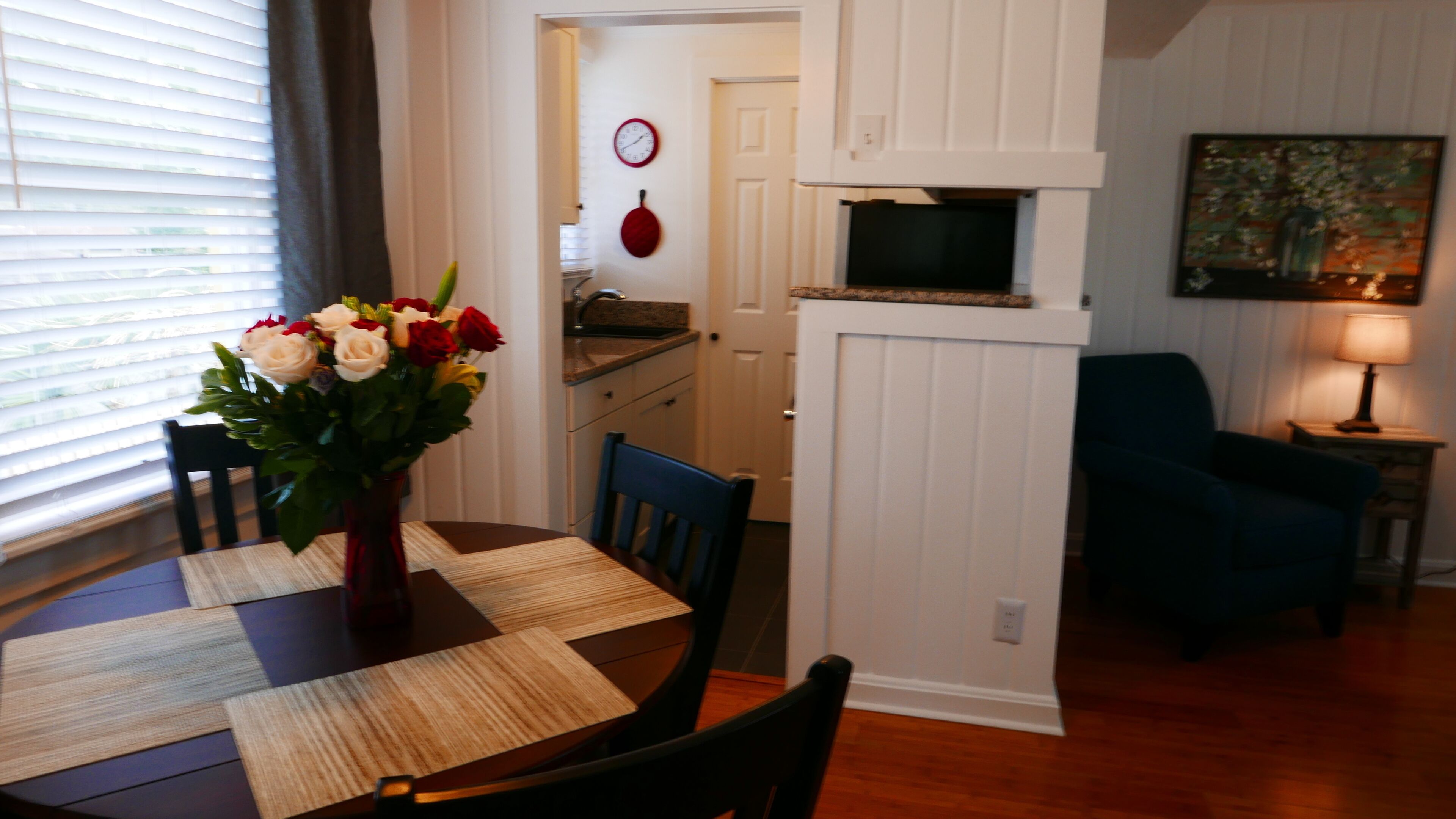 Un coin repas avec une table dressée pour deux personnes, un vase de fleurs et une cuisine en arrière-plan.