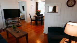 Uma sala de estar com televisão sobre um móvel, um sofá, uma mesa com abajur e um espaço de jantar com uma cadeira e um vaso de flores.