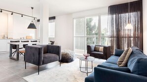 A modern living room with a dark blue sofa, a round coffee table, and a dining area with a white table and chairs. - Modern 2BR Apt w Private Parking in City Center (Ramat HaSharon)