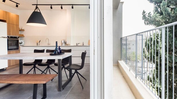 A modern kitchen with a dining area, a large window, and a balcony with a railing. - Modern 2BR Apt w Private Parking in City Center (Ramat HaSharon)