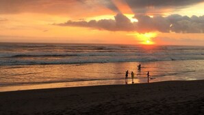 Une scène de plage avec un coucher de soleil, des vagues et quelques personnes debout sur le rivage.