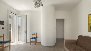 Un salon moderne avec un canapé beige, une chaise en bois et un fauteuil bleu. Une grande colonne blanche se trouve au centre, et une porte donne accès à une autre pièce.