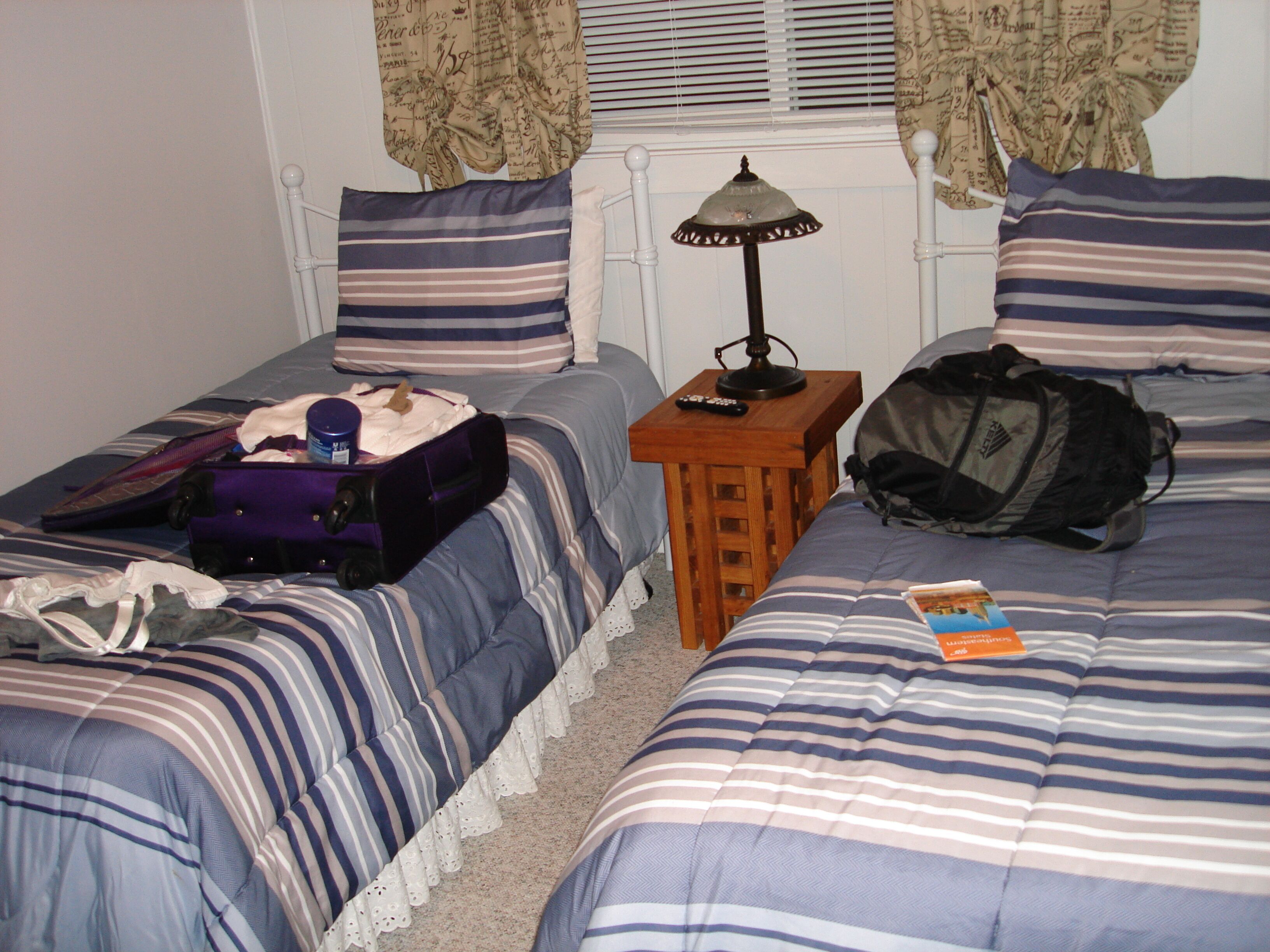 Una habitación con dos camas, cada una con ropa de cama y almohadas a rayas, una mesita de noche de madera con una lámpara, y una maleta sobre una cama.