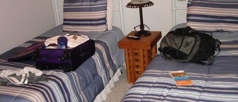Una habitación con dos camas, cada una con ropa de cama y almohadas a rayas, una mesita de noche de madera con una lámpara, y una maleta sobre una cama.