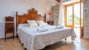 Una habitació amb un llit de fusta, roba de llit blanca, una tauleta de nit, una làmpada i vista a l'exterior.