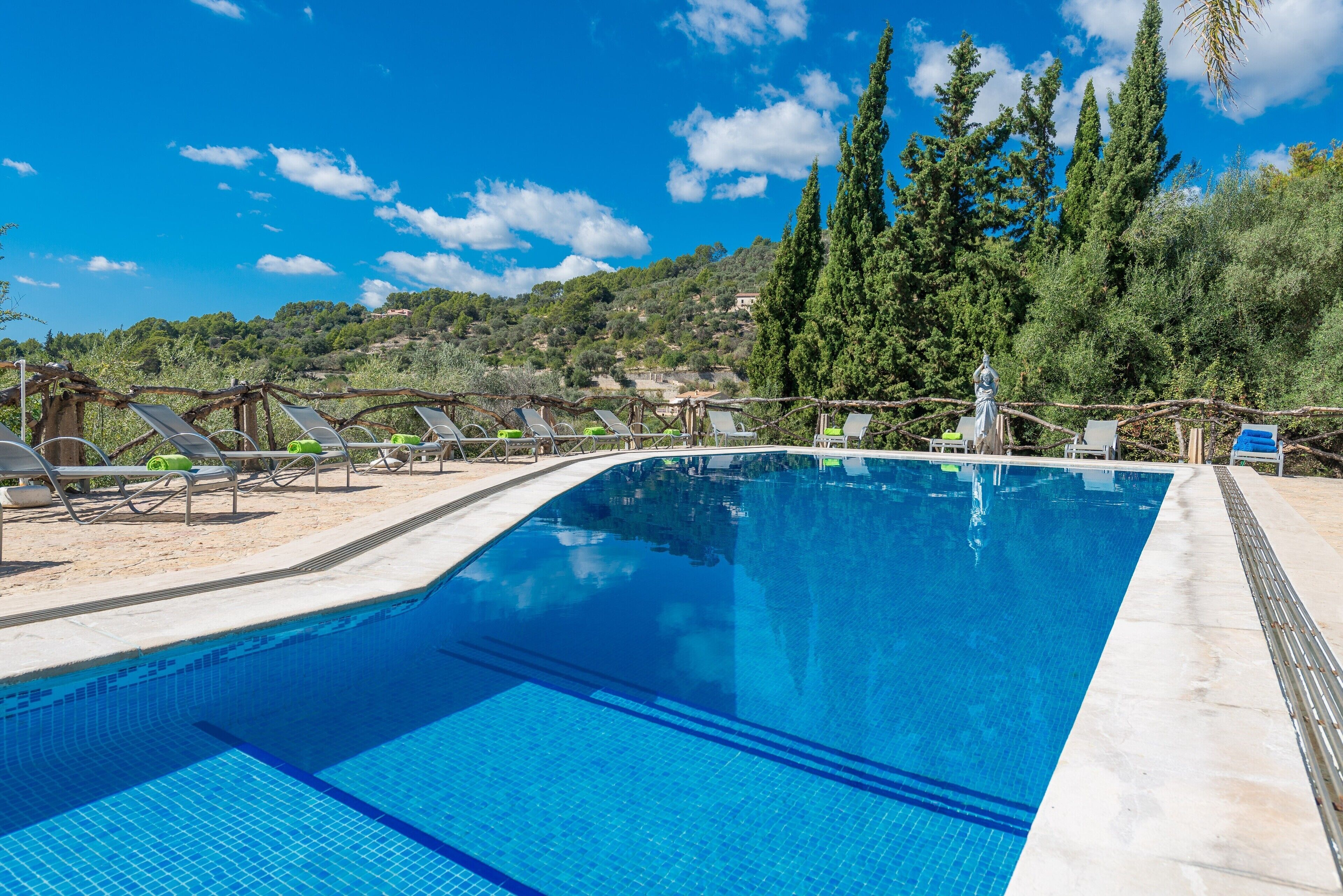 A clear blue swimming pool with lounge chairs and a statue, surrounded by trees and hills.