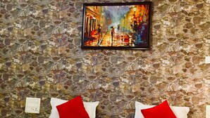 Habitación de hotel con pared de piedra, un cuadro de una escena callejera, dos almohadas rojas y una cama con manta roja.