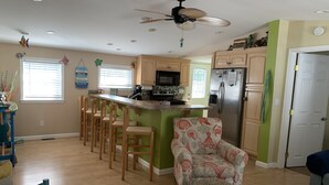A modern kitchen with a central island, wooden bar stools, a patterned armchair, and a ceiling fan. - SNOW BIRDS..SPECIAL RATES FOR WINTER TRAVELS - A Fun, Sun N' Surfin' Kinda Place (Murrells Inlet)