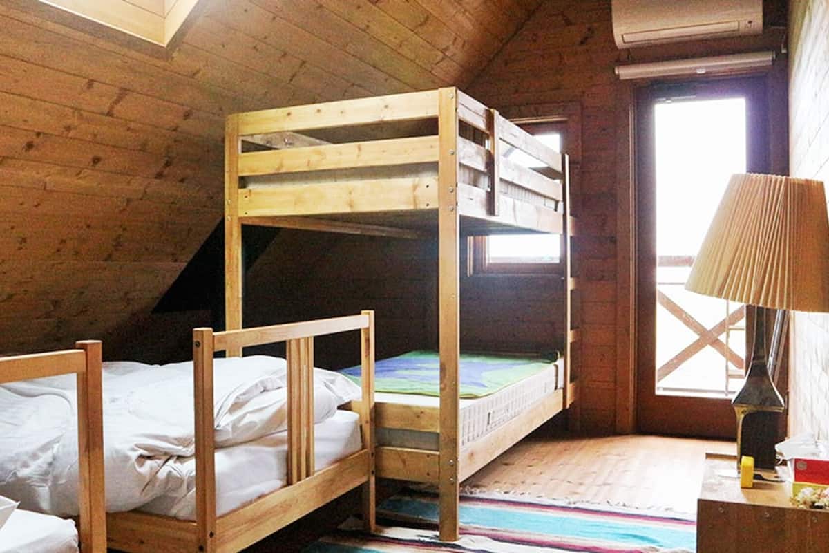 A dormitory room with two bunk beds, a wooden ceiling, a window, and a lamp on a bedside table.
