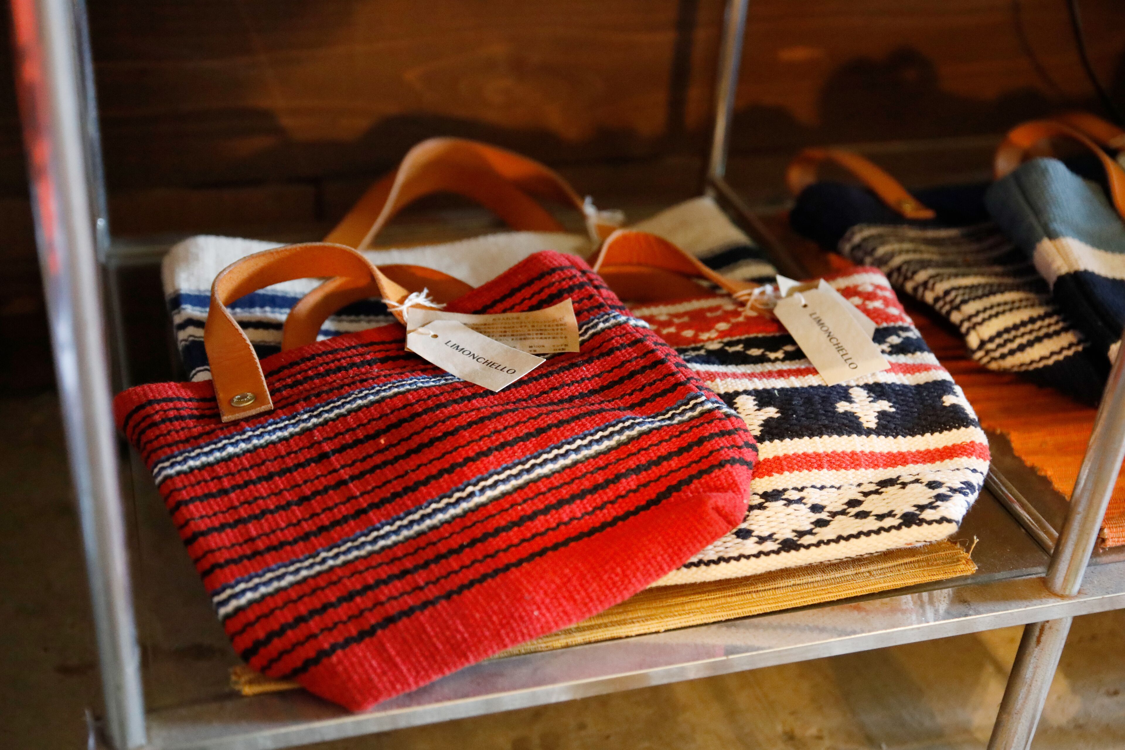 Une collection de sacs tissés, chacun muni d’une étiquette, est exposée sur une étagère en métal.