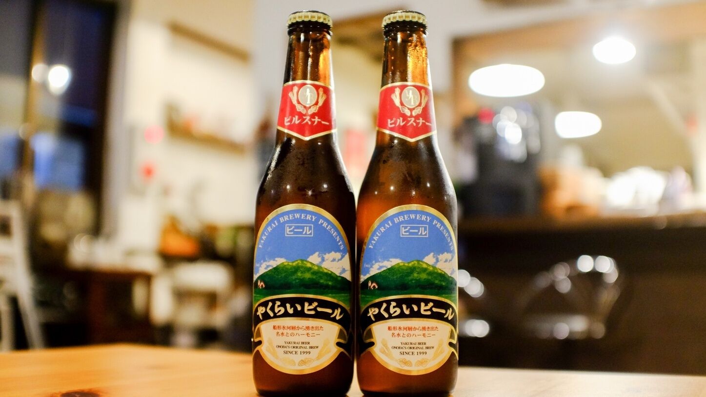 Two bottles of beer with labels, placed on a wooden surface in a setting with a blurred background.