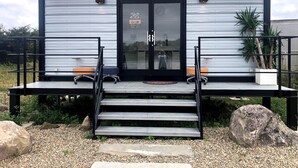 Un edificio moderno con fachada metálica, puerta de marco negro y un pequeño porche con escalones que dan a una zona de estar.