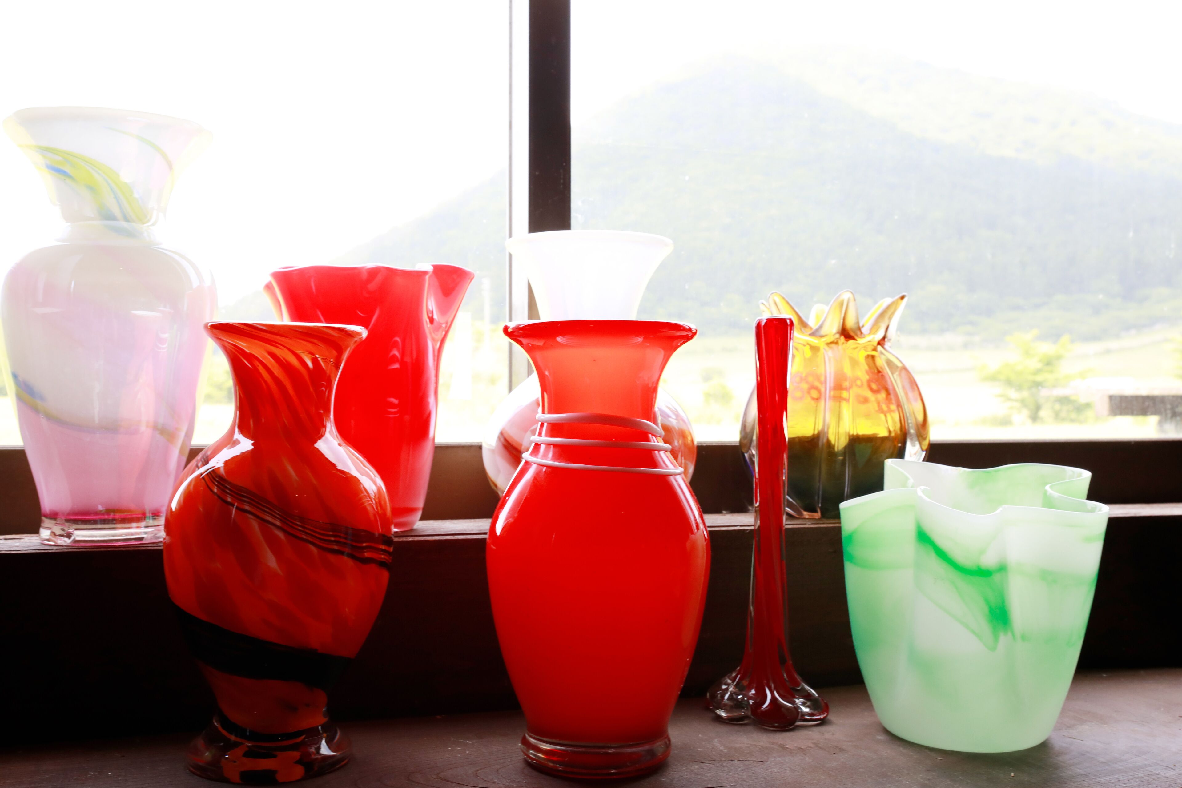 Un conjunto de coloridos floreros de vidrio sobre un alféizar con vistas a la montaña.