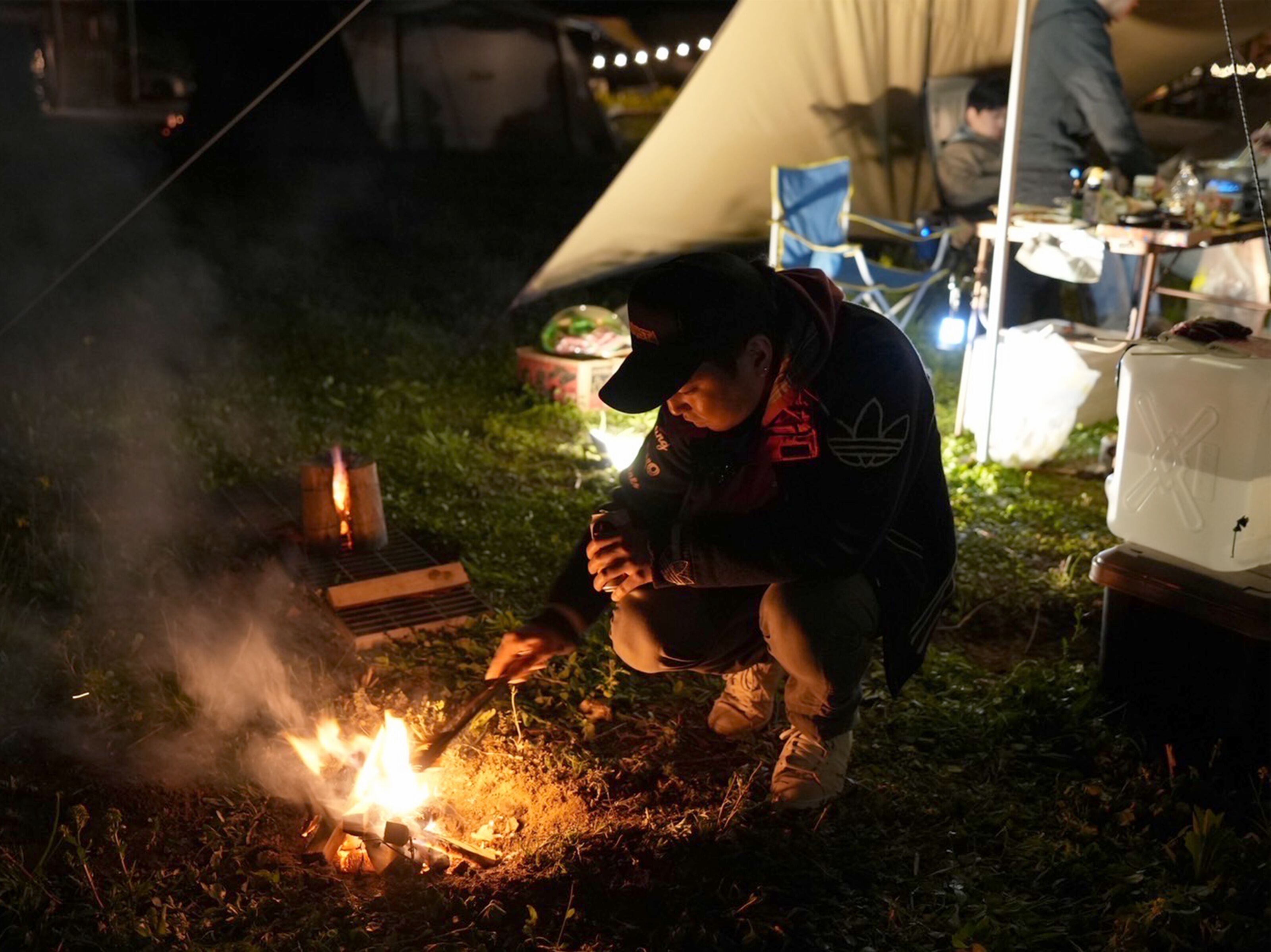 A person cooking outdoors at night with a campfire, surrounded by tents and camping equipment.