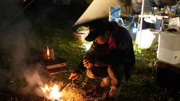 A person cooking outdoors at night with a campfire, surrounded by tents and camping equipment.
