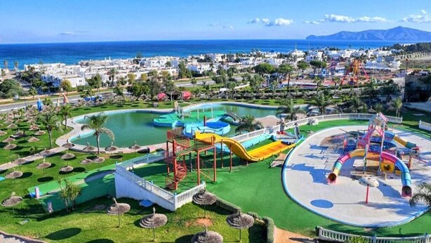 Een badplaats aan de kust met een groot waterpark, inclusief glijbanen en zwembaden, en uitzicht op zee en bergen op de achtergrond.