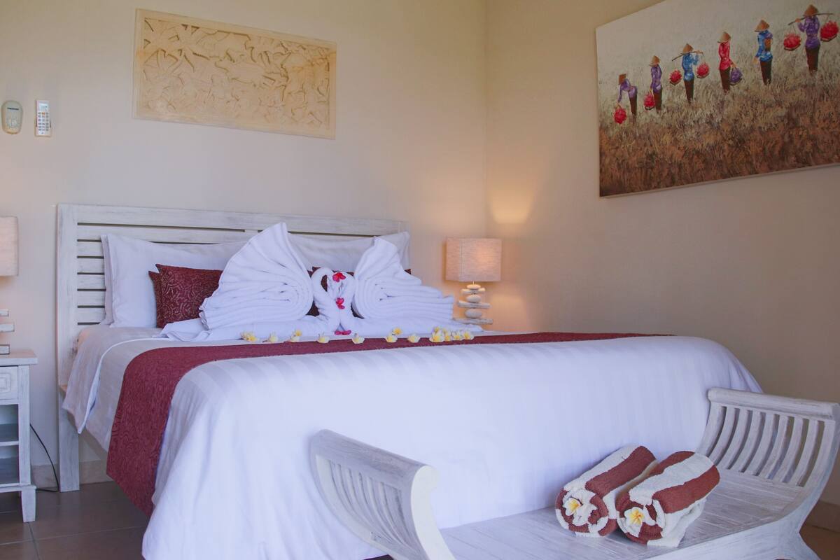 A neatly made bed with white linens and red accents, a bedside table with a lamp, and a painting of women in traditional attire on the wall.